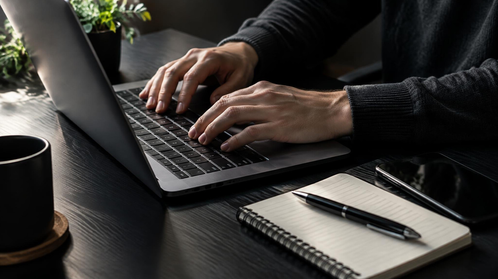Close-up of hands typing on a laptop next to a notebook and phone